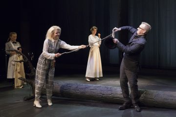 Ruudullinen nainen vetää lavalla pukumieheltä pitkää köyttä, ja kaksi vaaleisiin mekkoihin pukeutunutta naista seisoo heidän takanaan. Lattialla makaa suuri kaatunut puu rekvisiitta, ja taustalla on tumma sininen valaistus.