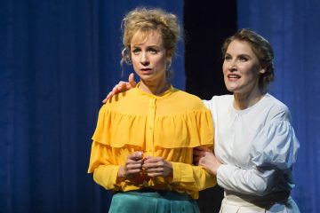 Lavalla seisoo kaksi naista; toinen kirkkaan keltaiseen puseroon pukeutunut näyttää huolestuneelta, kun taas toinen valkoisiin pukeutunut nainen hymyilee lempeästi ja rauhoittelee häntä kainalossaan. Taustalla roikkuvat siniset verhot.