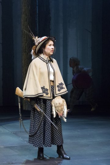 Lavalla seisoo nainen, jolla on pilkkihame, saappaat ja höyhenpäähattu, kädessään haulikko ja pehmolelu. Hänellä on koristeellinen viitta. Taustalla juoksee toinen henkilö, jolla on kädessään suuri punainen esine.