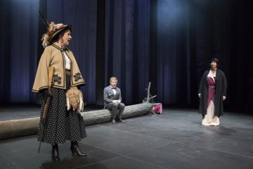 Kolme aikakauden pukuihin pukeutunutta näyttelijää esiintyy lavalla, jonka taustalla on pimeä metsä. Yksi nainen seisoo vasemmalla hattu ja viitta yllään, mies istuu kaatuneen hirren päällä keskellä, ja toinen nainen seisoo oikealla pitkässä mekossa ja takissa.