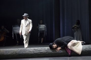 Vaaleaan pukuun ja hattuun pukeutunut mies seisoo näyttämöllä tummiin vaatteisiin pukeutuneen naisen vieressä, joka makaa tukin päällä, kun muut esiintyjät rekvisiittaineen ilmestyvät hämärään taustaan.