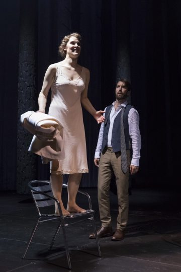 Vaaleaan slipsiin pukeutunut nainen seisoo paljain jaloin metallituolilla takki kädessään, kun liiviin ja pukupaitaan pukeutunut mies katsoo häntä. He ovat hämärästi valaistulla näyttämöllä, jossa on tummat verhot ja dramaattinen tunnelma.