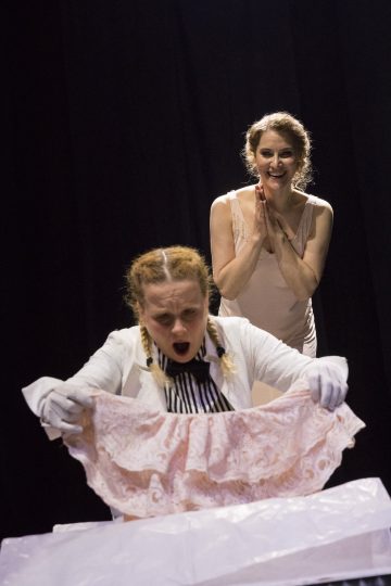 Kaksi naista näyttämöllä; toinen etualalla näyttää järkyttyneeltä ja pitelee vaaleanpunaista pitsistä vaatetta, kun taas toinen seisoo hänen takanaan hymyillen ja kädet jännittyneinä yhteen lyötynä. Molemmat ovat teatterivalaistuksen alla.