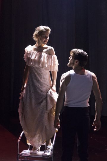 Nainen, jolla on pitsinen olkaimeton mekko, seisoo pienillä tikkailla ja katsoo alaspäin valkoiseen toppiin ja tummiin housuihin pukeutunutta miestä, joka katsoo häntä ylöspäin. Molemmat ovat dramaattisesti valaistuina hämärästi valaistulla näyttämöllä.