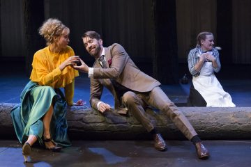 Lavalla esiintyy kolme näyttelijää; keltaiseen toppiin ja vihreään hameeseen pukeutunut nainen istuu tukin päällä ja jakaa juomaa ruskehtavaan pukuun pukeutuneen miehen kanssa, ja taustalla hymyilee ja halajaa polviaan kotiapulaisen asuun pukeutunut nainen.