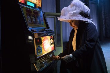 Henkilö, jolla on suuri, hienostunut valkoinen hattu ja tumma takki, seisoo peliautomaatin ääressä ja katsoo alaspäin, kun hän syöttää kolikoita hämärässä ympäristössä.