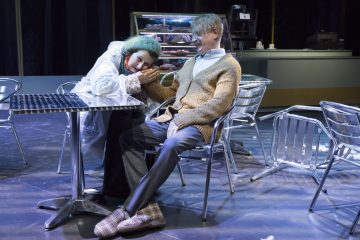 Kaksi teatteriasuihin pukeutunutta ihmistä istuu metallisen kahvilapöydän ääressä; toinen kumartuu hymyillen, vihreät hiukset ja klovnimeikki päässä, kun taas toinen, villatakki ja ruudulliset tossut jalassa, hymyilee takaisin. Taustalla on vitriini ja kahvilan tiski.