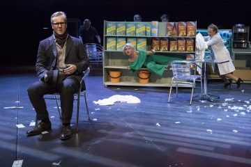 Pukumies istuu tuolilla lavalla, kun vihreäpukuinen nainen ryntää muropaketeilla varustettujen hyllyjen läpi. Kaksi muuta katselee, ja valkopukuinen nainen seisoo kahvilan pöydän ääressä. Lattialle on ripoteltu rahaa ja höyheniä.