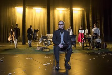 Mies istuu yksin tuolilla dramaattisen lavastuksen etualalla, ja hänen takanaan on viisi muuta ihmistä, soittimia ja rekvisiittaa lämpimän keltaisessa valaistuksessa ja metsää muistuttavan taustan alla.