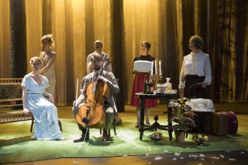 Kuuden henkilön ryhmä vintage-vaatteissa poseeraa metsää muistuttavan lavastuksen päällä. Yksi mies istuu ja soittaa selloa, kun muut seisovat tai istuvat lähellä, ja etualalla on pöytä, jossa on kakku, kynttilöitä ja juomia.