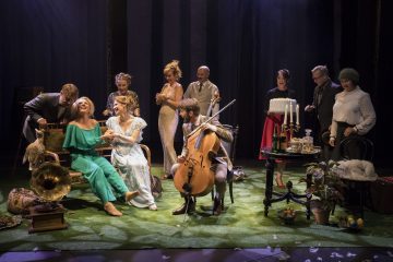 Joukko tyylikkäisiin pukuihin pukeutuneita ihmisiä on kokoontunut lavalle, jossa on vihreää ruohoa, soittimia ja vintage-koristeita, mikä viittaa teatteriesitykseen tai harjoituksiin, joissa on vilkasta ja iloista vuorovaikutusta.