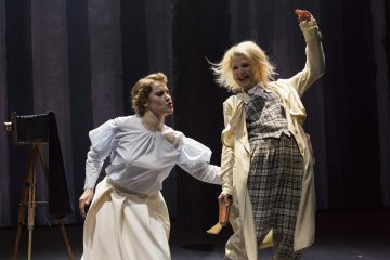 Lavalla on kaksi näyttelijää, joista toisella on valkoinen pusero ja hame, ja hän ojentaa kätensä, kun taas toinen, jolla on ruudullinen puku ja pitkä takki, pitää kättä ylhäällä. Taustalla seisoo vanhanaikainen kamera.