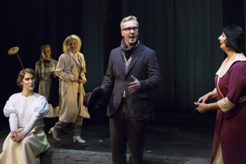 Lavalla puhuu intohimoisesti pukuun ja huiviin pukeutunut mies, jolla on hattu kädessään. Neljä aikakauden pukuihin pukeutunutta naista kuuntelee; kaksi seisoo taustalla, yksi istuu valkoisessa, ja toinen seisoo etualalla violettiin ja mustaan pukeutuneena. Teatteriympäristö.