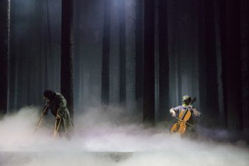 Kaksi esiintyjää sumuisella näyttämöllä, joka on lavastettu pimeäksi metsäksi; toinen soittaa selloa, toinen kävelee kainalosauvojen kanssa, molemmat osittain sumun ja korkeiden puiden läpi suodattuvan hämärän valon peittäminä.