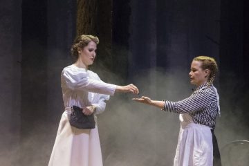 Kaksi naista seisoo vastakkain hämärästi valaistulla näyttämöllä. Toinen valkoiseen mekkoon pukeutunut nainen ojentaa jotain toiselle naiselle, jolla on raidallinen toppi ja valkoinen esiliina. Tausta on sumuinen ja siinä on korkeita, tummia pylväitä.