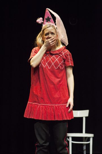 Punamekkoon ja vaaleanpunaiseen juhlahattuun pukeutunut nainen seisoo lavalla ja peittää suunsa toisella kädellään ja näyttää yllättyneeltä tai järkyttyneeltä. Taustalla näkyy valkoinen tuoli tummaa taustaa vasten.