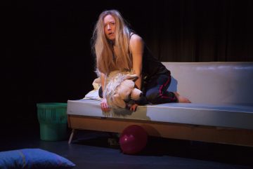 Mustiin vaatteisiin pukeutunut, pitkät vaaleat hiukset omaava nainen istuu sohvalla pitelemässä suurta pehmoeläintä. Lattialla on punainen ilmapallo, ja hänen vieressään on vihreä roskakori ja tyyny. Tausta on tumma ja dramaattinen.