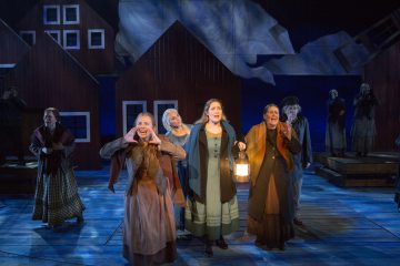 Lavalla seisoo joukko aikakauden asuihin pukeutuneita naisia, joista osa huutaa tai laulaa, ja yhdellä on sytytetty lyhty kädessään. Puiset talot ja dramaattinen valaistus luovat taustalle intensiivisen, teatterillisen tunnelman.