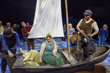 Ryhmä aikakauden pukuihin pukeutuneita näyttelijöitä esiintyy lavalla. Yksi vihreäpukuinen nainen istuu pienessä puisessa purjeveneessä muiden ympäröimänä, kun he näyttelevät vilkasta kohtausta näyttämön valojen alla.