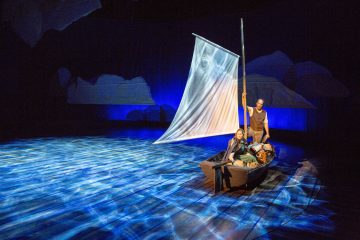 Kaksi näyttelijää esiintyy näyttämöllä pienessä veneessä, jossa on valkoinen purje ja jota ympäröi vettä jäljittelevä sininen valaistus. Toinen istuu, toinen seisoo, molemmat dramaattisessa valaistuksessa, ja taustalla on jäävuoria muistuttavia abstrakteja muotoja.