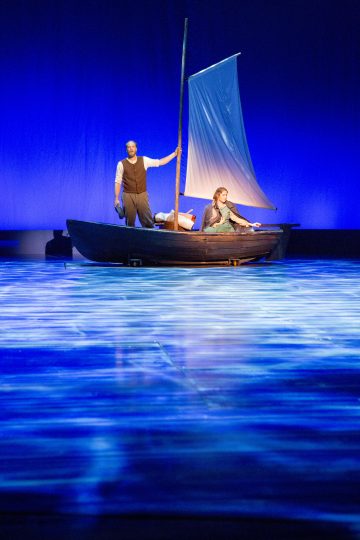 Kaksi ihmistä pienessä purjeveneessä on lavalla sinisessä valaistuksessa, joka antaa vaikutelman, että he kelluvat veden päällä, ja toinen seisoo ja pitää mastoa ja toinen istuu ja katsoo ulos.