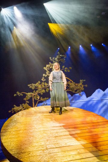 Vaaleansiniseen mekkoon ja silmälaseihin pukeutunut nainen seisoo pyöreällä, puisella näyttämöllä dramaattisessa valaistuksessa, ja taustalla näkyy puu ja sinisiä vuoren muotoja.