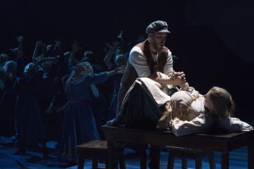 Mies seisoo pöydällä makaavan naisen yläpuolella pitäen tätä käsistä kiinni, kun heidän takanaan oleva ryhmä ihmisiä ojentaa kätensä dramaattisin elein hämärässä, teatterimaisessa valaistuksessa.