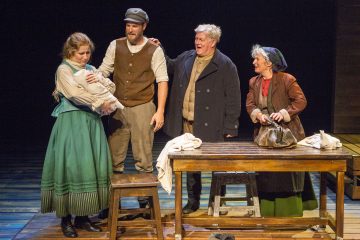 Neljä aikakauden pukuihin pukeutunutta näyttelijää seisoo näyttämöllä puisen pöydän ympärillä; yksi nainen pitelee vauvaa, kun muut hymyilevät ja elehtivät lämpimästi, mikä viittaa perhekohtaukseen teatterituotannossa.