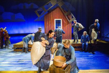 Näyttelijät esiintyvät näyttämöllä kallistetun punaisen talon edessä sinisissä puvuissa ja kantavat rekvisiittaa, kuten säkkejä ja puisia kulhoja, ja sinisellä valaistulla taustalla on ulkoilmakuvaan viittaava tausta.