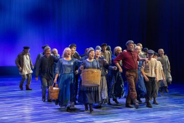 Ryhmä aikakauden pukuihin pukeutuneita näyttelijöitä esiintyy energisesti lavalla, ja joillakin on kädessään puisia ämpäreitä. Taustalla on sinisiä valoja, jotka luovat dramaattisen teatteritunnelman.