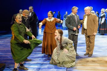 Ryhmä aikakauden pukuihin pukeutuneita näyttelijöitä esiintyy lavalla. Yksi vihreäpukuinen nainen elehtii dramaattisesti, kun taas toinen ruutupukuinen nainen kyykistyy ja huutaa. Muut reagoivat yllätyksellä tai naurulla sinistä taustaa vasten.