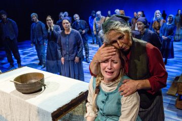 Vanhempi nainen lohduttaa itkevää nuorta naista, joka on pukeutunut aikakauden vaatteisiin ja jota ympäröi joukko ihmisiä hämärästi valaistulla näyttämöllä, mikä viittaa teatteriesityksen tunteikkaaseen kohtaukseen.