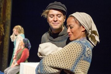 Tummahattupäinen mies hymyilee huivipäisen naisen vieressä, jolla on niputettu vauva kädessään. Taustalla lavalla istuu kaksi muuta, hieman epäselvää ihmistä, kaikki lämpimissä, maalaismaisissa vaatteissa.