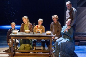 Kuusi vanhanaikaisiin vaatteisiin pukeutunutta ihmistä kokoontuu maalaismaisen puupöydän ympärille, jossa on kulhoja ja yksi kynttilä. Kohtaus vaikuttaa teatterimaiselta, ja siinä on dramaattinen valaistus ja tumma näyttämötausta.