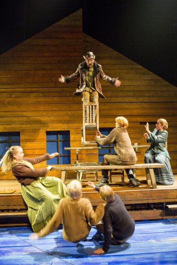 Mies seisoo näyttämöllä pöydän päällä olevalla tuolilla kädet ojennettuina, ja viisi naista pöydän ympärillä reagoi dramaattisesti. Tausta muistuttaa puutaloa.