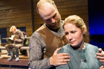 Parrakas mies pitelee hellästi naisen olkapäitä, kun tämä katsoo alaspäin huolestuneena; taustalla toinen mies istuu penkillä lukemassa kirjaa. Kohtaus näyttää olevan näyttämönäytelmästä, jossa on dramaattinen valaistus.