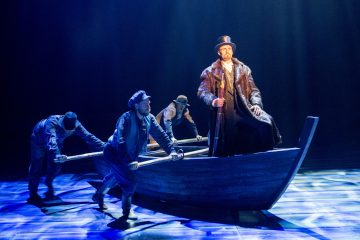 Neljä näyttelijää esiintyy näyttämöllä dramaattisessa valokeilassa. Kolme työläisiksi pukeutunutta työntää suurta puuveneen rekvisiittaa, kun taas neljäs, turkistakkiin ja silinterihattuun pukeutunut näyttelijä istuu veneessä keppi kädessään.