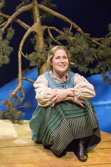 Aikakauden asuun pukeutunut nainen, jolla on vihreä mekko ja raidallinen hame, istuu hymyillen puisella näyttämöllä, kädet polviensa ympärillä. Hänen takanaan on mänty ja sininen tausta, jotka viittaavat ulkokohtaukseen.