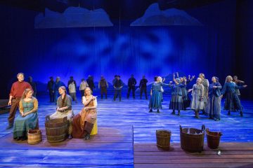 Näyttämöllä nähdään suuri joukko aikakauden pukuihin pukeutuneita näyttelijöitä, joista osa istuu etualalla olevien puutynnyreiden ääressä ja osa seisoo ryhmissä. Lavastus on valaistu sinisellä valolla, ja yläpuolella roikkuu pilviä.