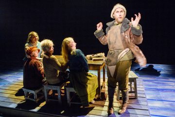 Maalaismaisesti vanhanaikaisiin vaatteisiin pukeutunut mies esiintyy lavalla eloisasti neljän puupöydän ääressä istuvan lapsen edessä, jotka katselevat häntä kiinnostuneina ja yllättyneinä kirkkaiden näyttämövalojen alla.