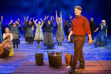 Hymyilevä mies, jolla on punainen paita ja lippis, seisoo näyttämön keskellä sipulia kädessään, puuämpäreitä jalkojensa juuressa. Hänen takanaan joukko vanhanaikaisiin vaatteisiin pukeutuneita ihmisiä nostaa käsiään koreografioidun tanssin tahdissa puisella lavalla, jossa on sininen valaistus.