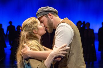 Mies ja nainen puvussa jakavat suudelman lavalla dramaattisen sinisellä valaistulla taustalla ja muiden esiintyjien silueteilla kaukaisuudessa.