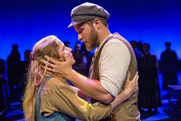 Aikakauden vaatteisiin pukeutuneet mies ja nainen syleilevät läheisesti näyttämöllä katsoen toistensa silmiin, ja heidän takanaan on syvän sininen tausta ja muiden ihmisten siluetteja.