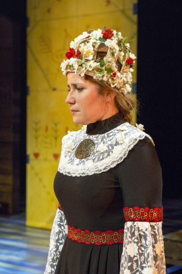 Profiilikuva naisella on perinteinen puku, jossa on pitsisiä yksityiskohtia ja kukkaompeleita, sekä valkoisista ja punaisista kukista tehty kruunu. Hän seisoo sisätiloissa keltaista kuviota vasten ja katsoo mietteliäästi.