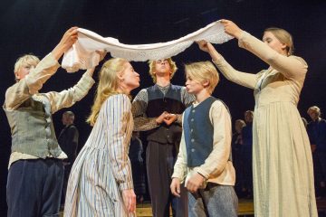 Viisi vanhanaikaisiin vaatteisiin pukeutunutta nuorta seisoo yhdessä lavalla. Kaksi heistä pitelee pitsireunuksin koristeltua valkoista liinaa toisiaan vastakkain seisovien pojan ja tytön päiden yläpuolella, ja toinen henkilö seisoo heidän takanaan.