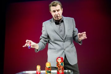 Harmaapukuinen mies elehtii ja pitää kädessään pientä venäläistä pesänukkea. Useita muita nukkeja on järjestetty hänen eteensä kirkkaalle pöydälle, ja taustalla on yhtenäinen punainen väri.