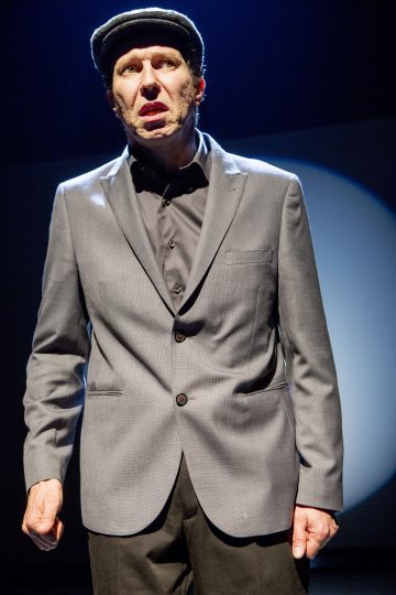 Harmaaseen pukuun ja mustaan litteään lippalakkiin pukeutunut mies seisoo lavalla valonheittimen alla ja katsoo intensiivisesti eteenpäin suu auki ja nyrkki kylkeään vasten.