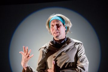 Henkilö, jolla on sininen päähine ja beige takki, tekee ilmeikkäitä eleitä puhuessaan ja seisoo tummalla taustalla olevan pyöreän valokeilan edessä.