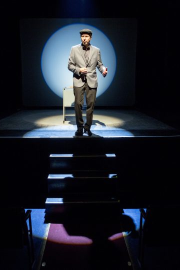 Pukuun ja hattuun pukeutunut mies seisoo yksin hämärässä, valonheittimen valaisemassa näyttämöllä, ja hänen takanaan on pieni pöytä ja etualalla tyhjiä teatteripenkkejä.