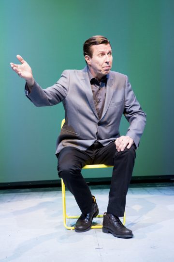 Harmaaseen bleiseriin ja mustiin housuihin pukeutunut mies istuu keltaisella tuolilla vihreää taustaa vasten ja elehtii vasemmalla kädellään ja ilmeikäs ilme kasvoillaan.